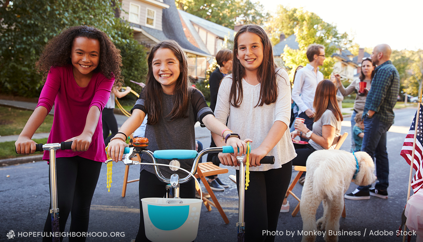 Photo shows three girls on scooters and bikes at a block party, adults and a white dog in the background.