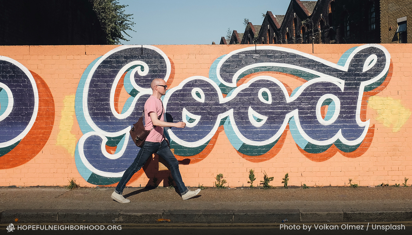 Young man walking on a sidewalk with a painted wall behind him. The painted wall says "Good" in cursive.