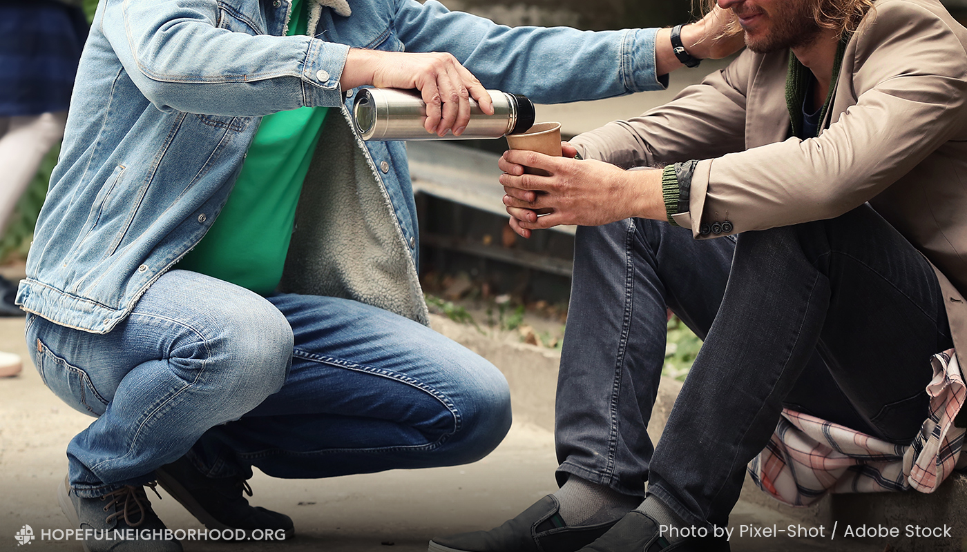 Photo of a man pouring coffee into the cup of a man sitting on the sidewalk.