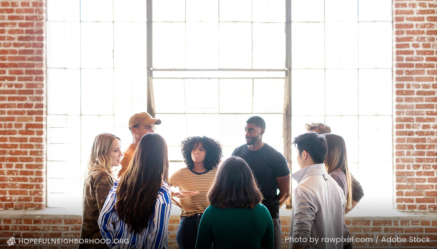 A group of young adults hanging out in a warehouse-type space with a large window behind them.