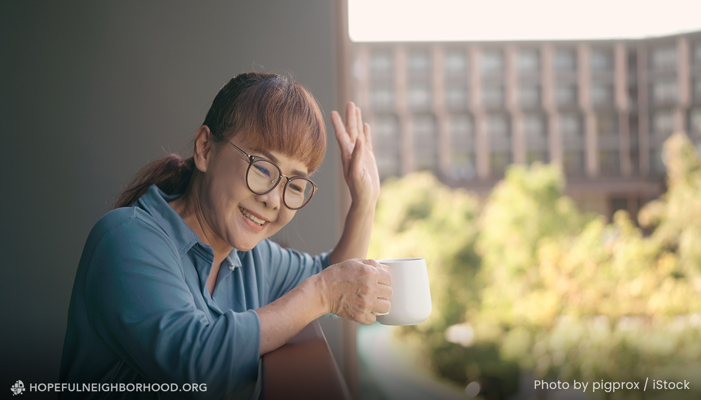 A woman is waving from her balcony while holding a cup of coffee.