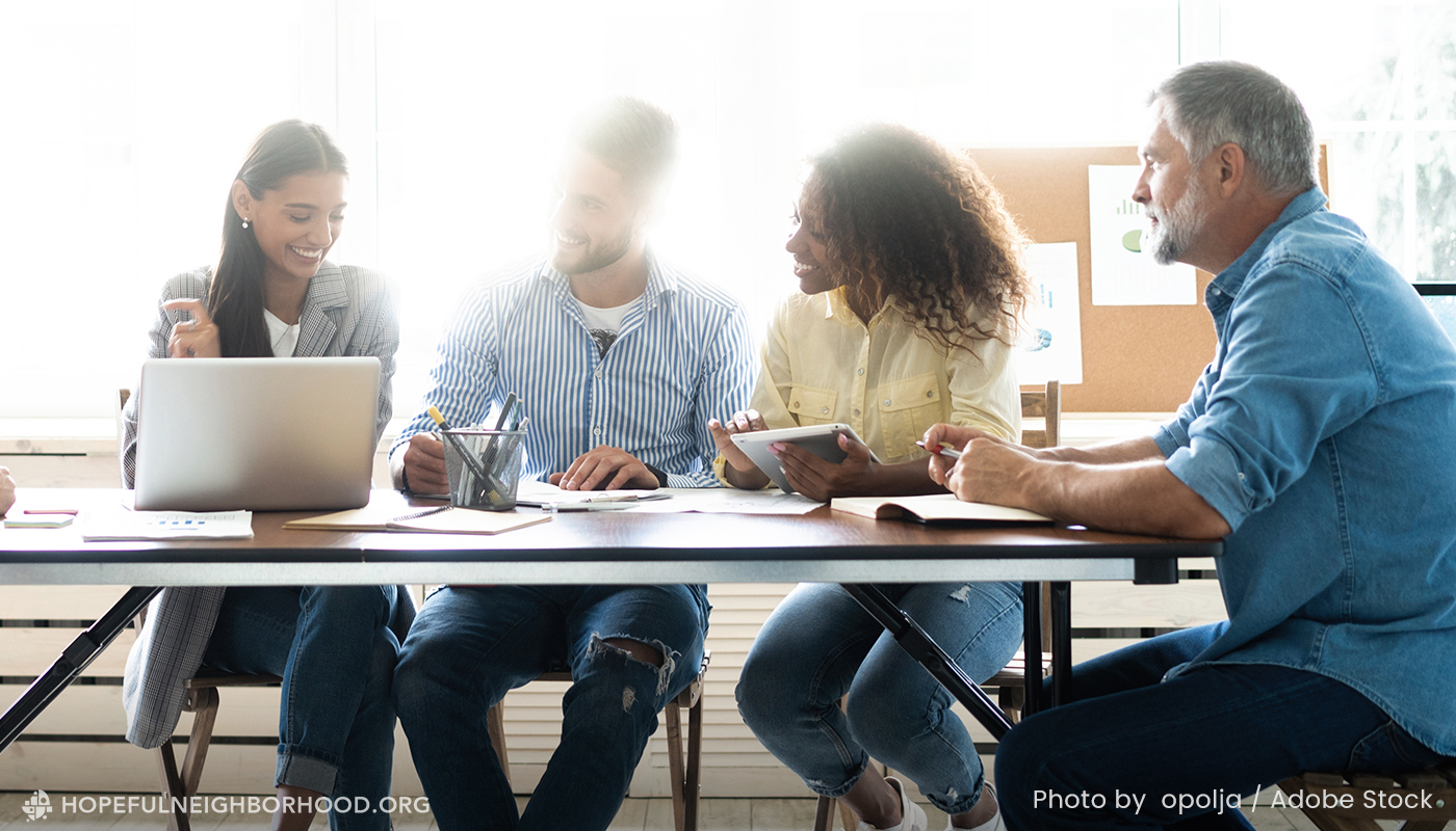 A community group is meeting at a table with bright sunshine coming through the window behind them.